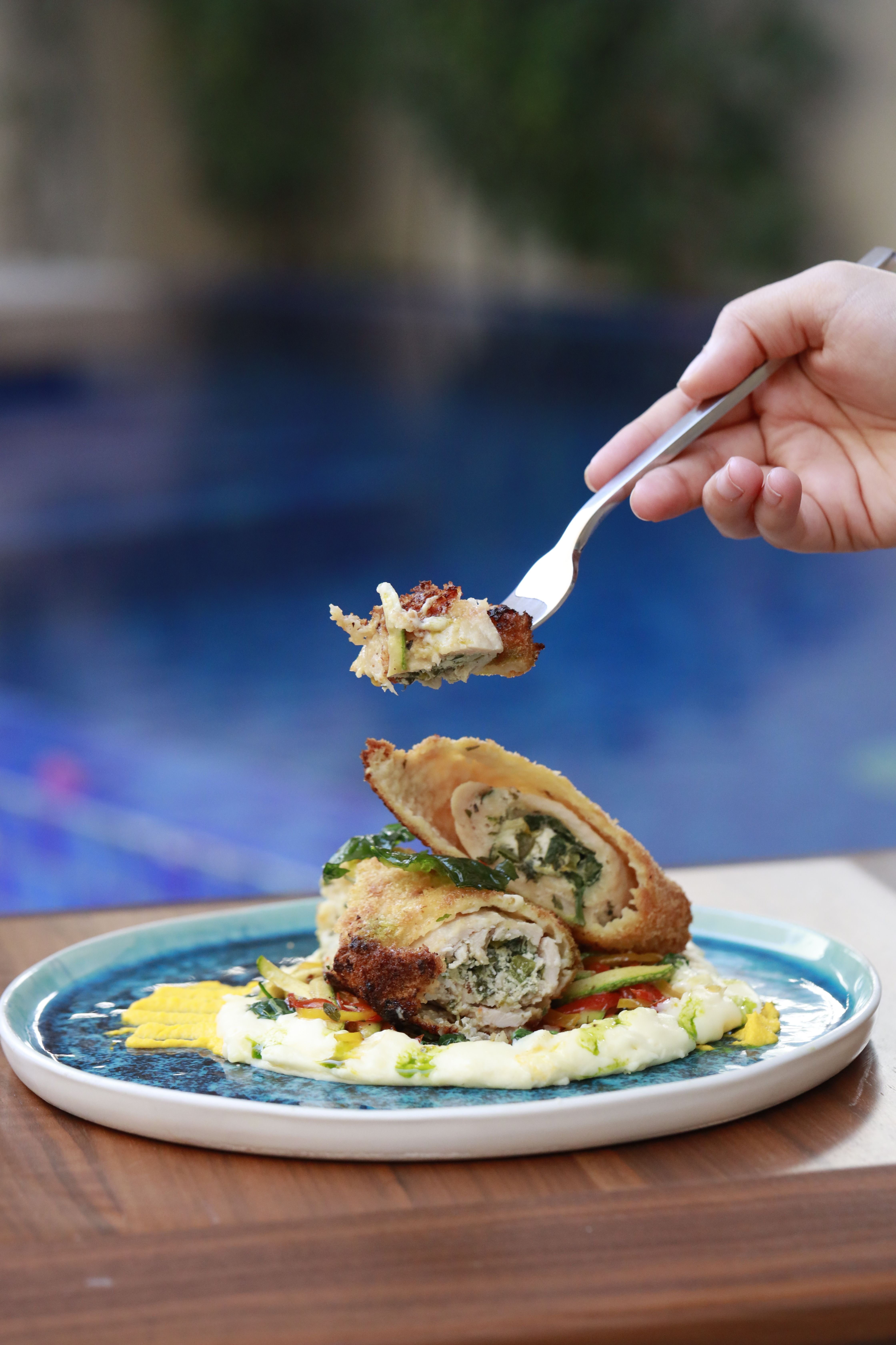 Close-up of a gourmet chicken meal being lifted by a fork over a blue plate outdoors.