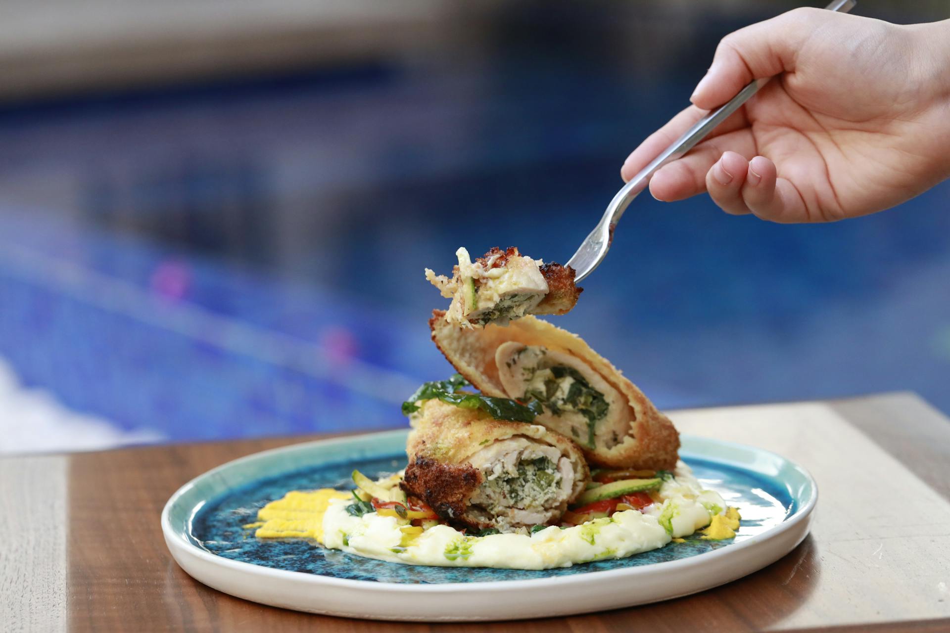 Close-up of a fork lifting a piece of chicken roulade on a plate with a poolside view.