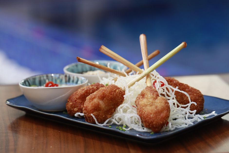 Golden-grilled shrimp paste molded around sugarcane sticks arranged on a platter with lettuce leaves, mint, and rice paper wrappers next to a small bowl of dipping sauce.