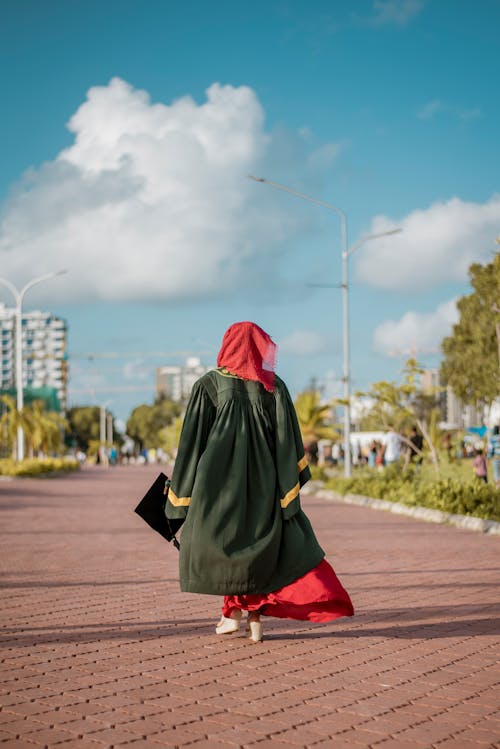 Back View of Woman in a Graduation Gown and Holding a Mortarboard Walking on a Sidewalk 