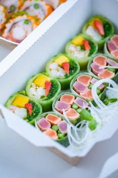 Vibrant sushi rolls with salmon, tuna, mango, and seaweed in a takeout box.