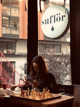 Woman using a mobile phone in a cozy café, with a chessboard on the table.