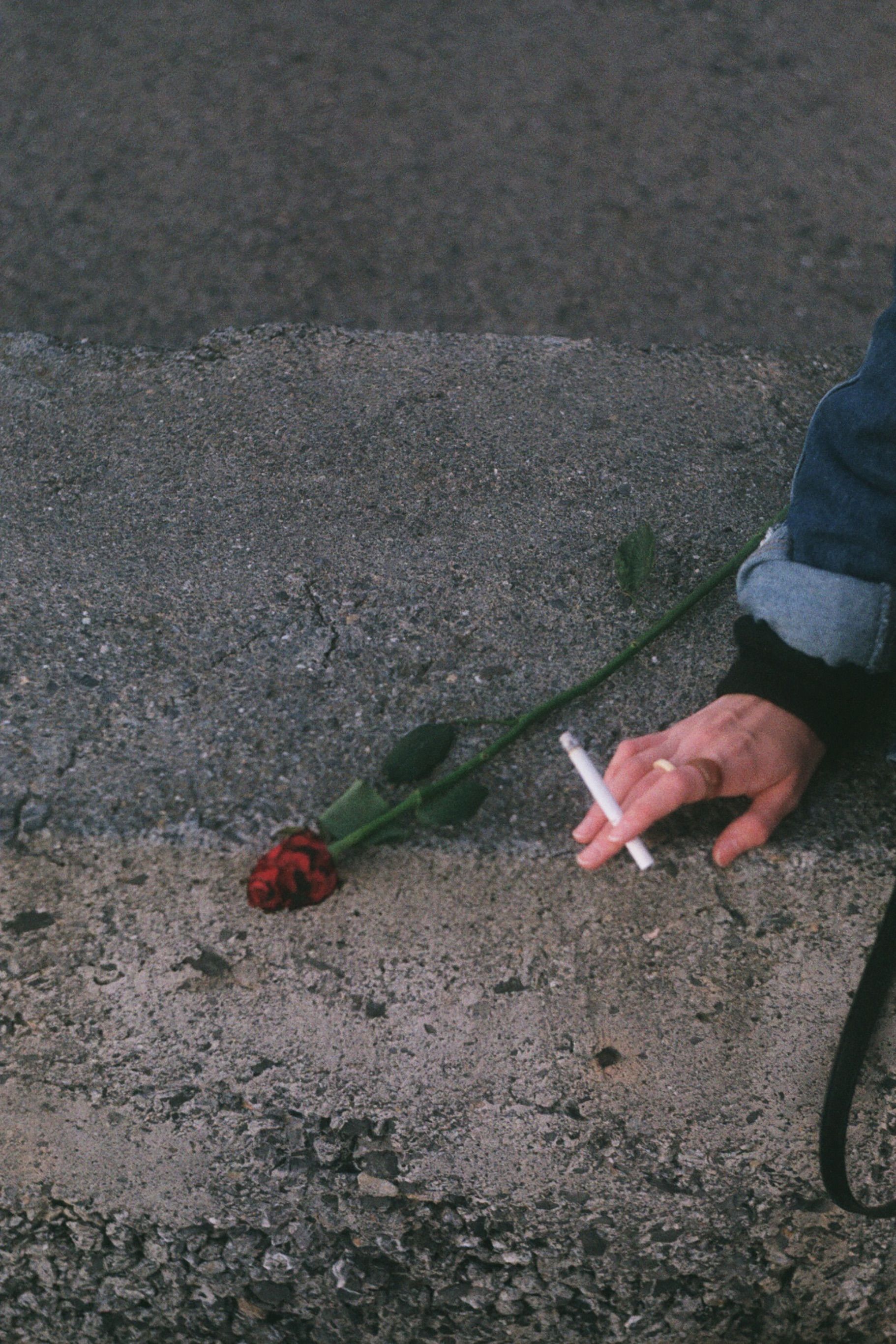 A person holding a cigarette beside a red rose on concrete outdoors.