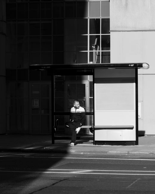 Monochrome photo of a man sitting at an urban bus stop, waiting.