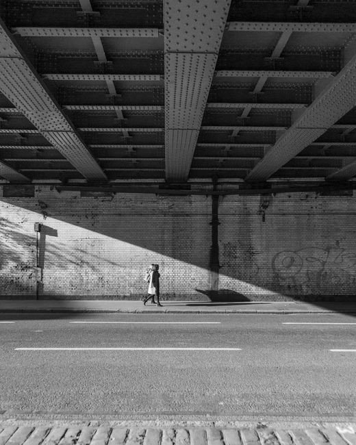 Monochrome image capturing a lone person walking under a steel bridge on a city street.