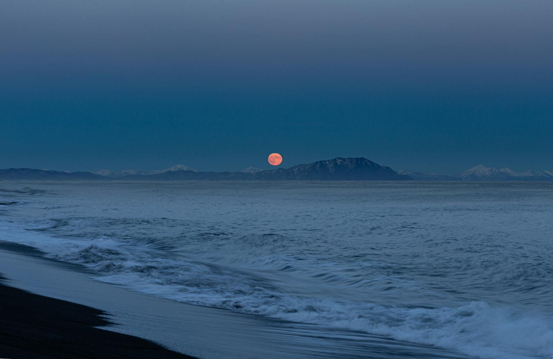 A serene beach with waves and a full moon over distant mountains at twilight.