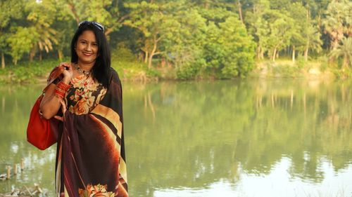Free A woman in a floral dress posing happily by a serene lake surrounded by greenery. Stock Photo