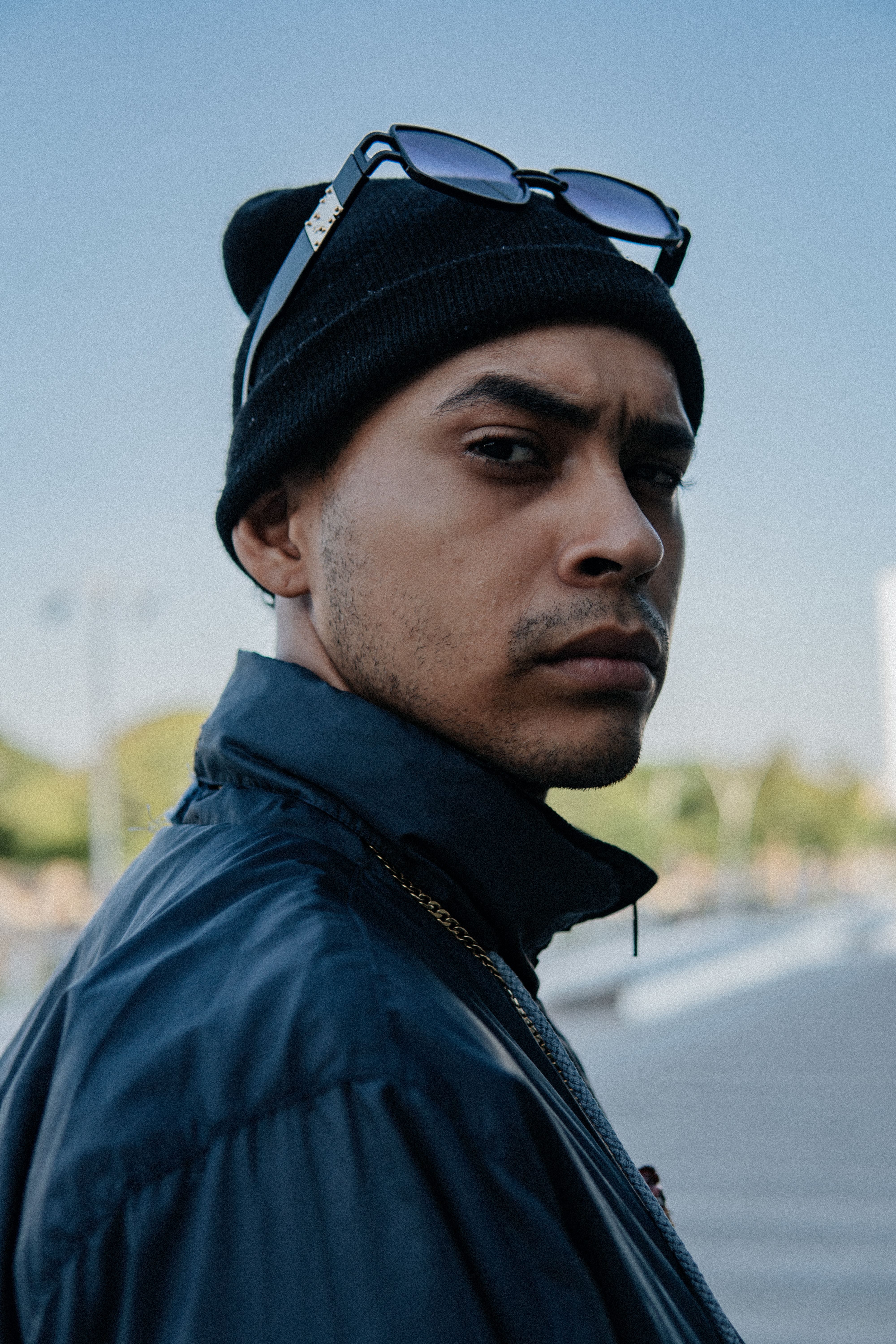 Portrait of a serious man in a beanie and sunglasses, outdoors with a defocused city background.
