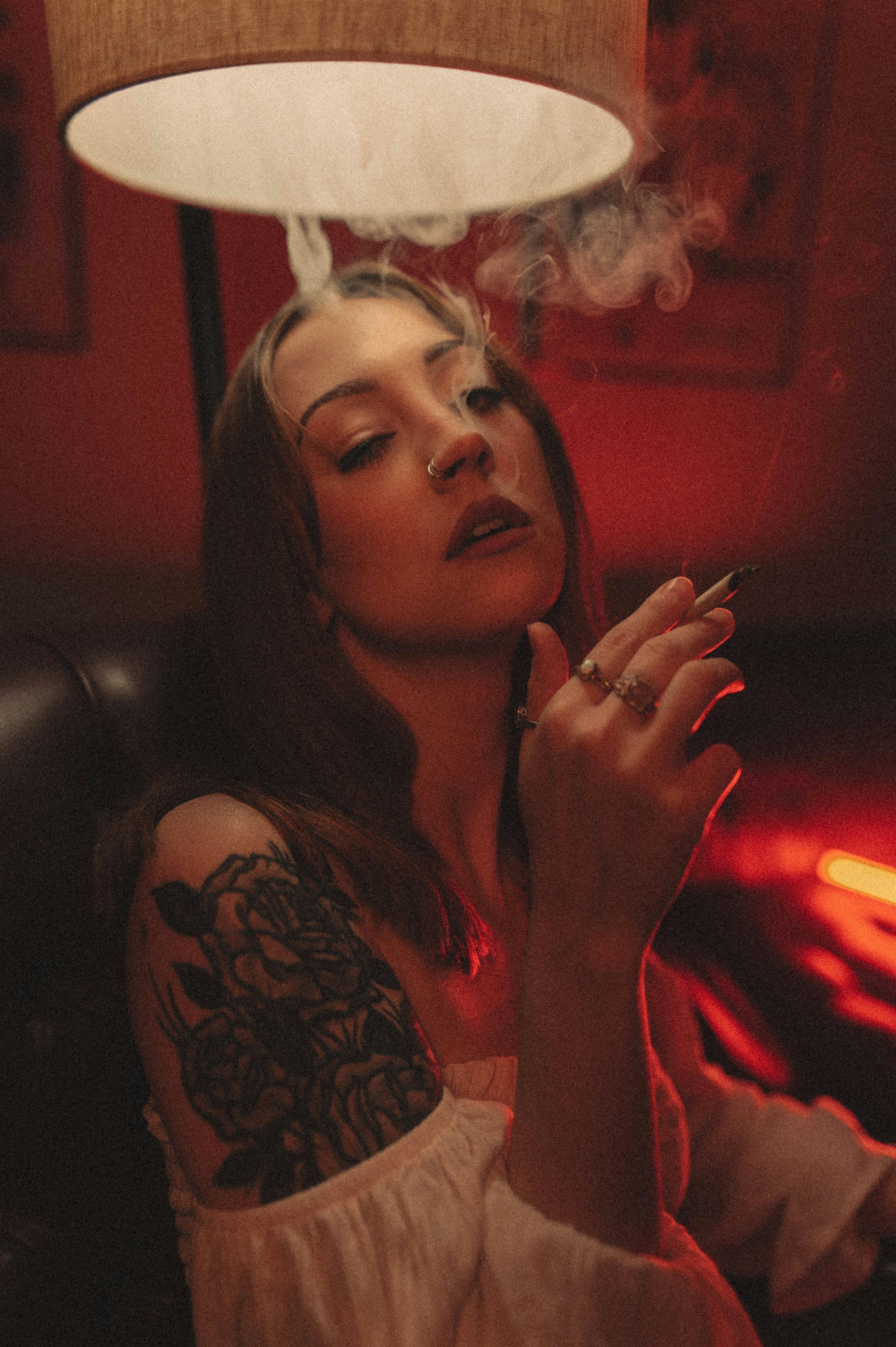 A tattooed woman smokes in a dimly lit lounge under warm light.