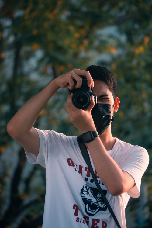 Free Man in a white shirt wearing a face mask, capturing photos outdoors with a camera. Stock Photo