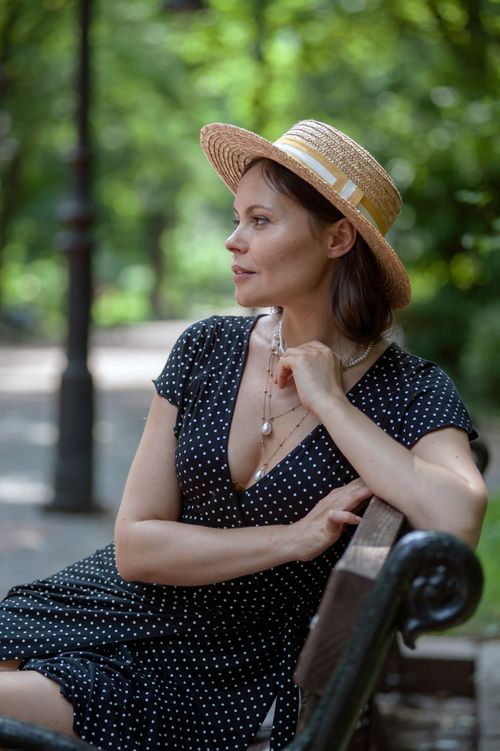 Free A woman wearing a polka dot dress and straw hat sits on a bench in a lush park, looking relaxed. Stock Photo