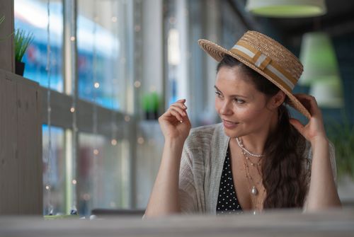 Free A fashionable woman in a straw hat smiles indoors at a chic urban cafe, exuding casual elegance. Stock Photo