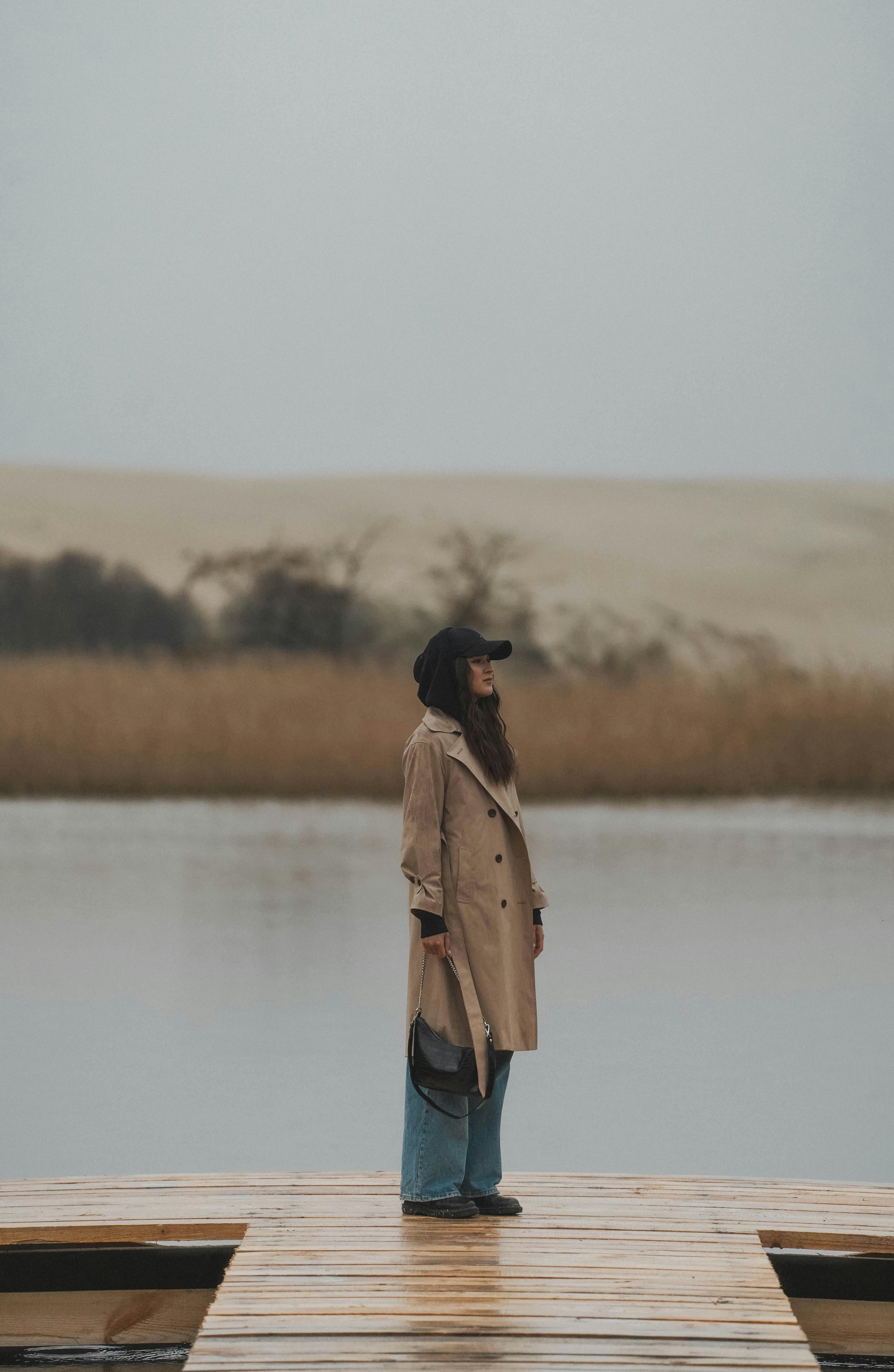 A woman in a trench coat stands on a wooden pier by a serene lake during autumn in Kaliningrad, Russia.