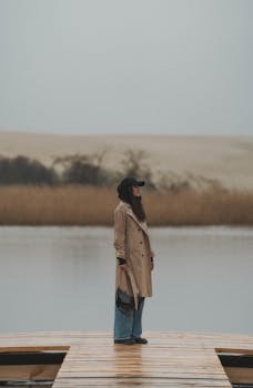 A woman in a trench coat stands on a wooden pier by a serene lake during autumn in Kaliningrad, Russia.