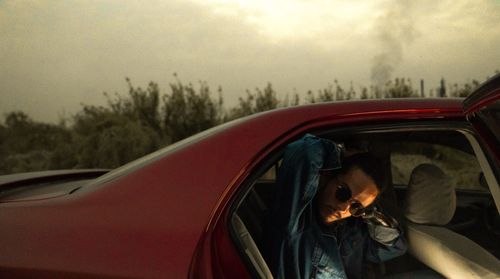 Free A man in sunglasses and denim jacket steps out of a red car at dusk in a rural setting. Stock Photo