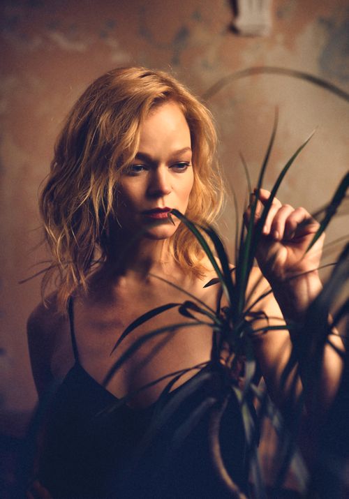 Free Thoughtful blonde woman in intimate setting, softly touching a houseplant in warm, moody lighting. Stock Photo