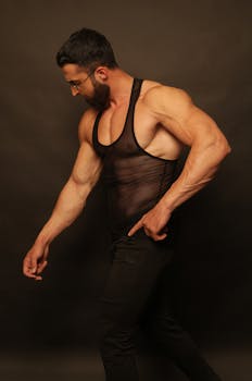 A muscular bodybuilder poses confidently in a dark studio wearing a see-through tank top.