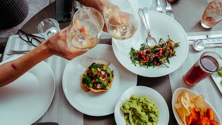 Two Unrecognizable Hand Holding Glasses Of Wine Above Plates Of Colourful Salads