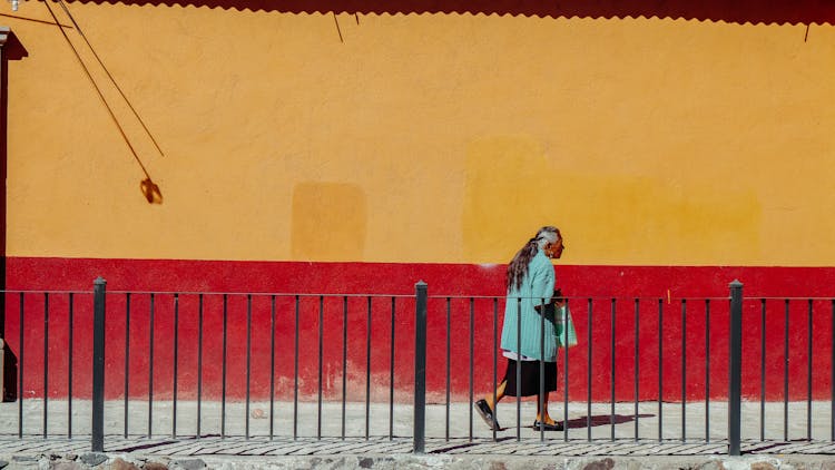 Old Woman Walking Down Street Along Red And Yellow Wall