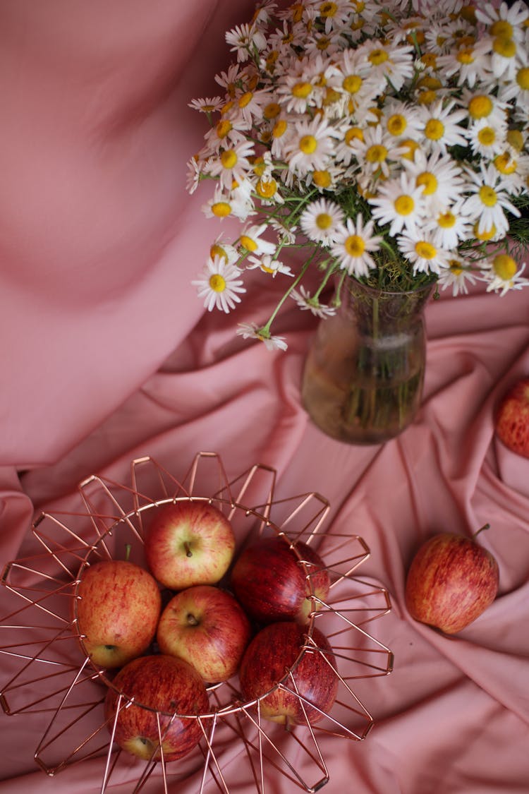 Red Apples In Metal Basket And Bunch Of Flowers Laying On Pink Tablecloth 