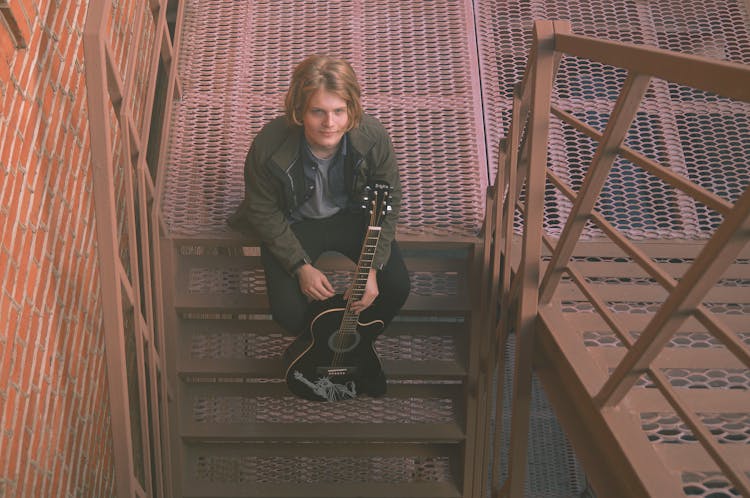 A Man Sitting On Steel Staircase Holding A Black Guitar
