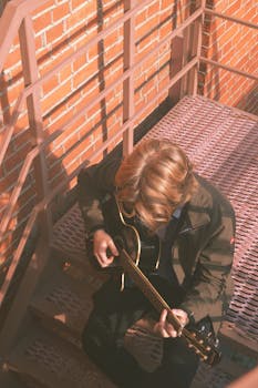 A man with blonde hair playing an acoustic guitar on outdoor stairs. Warm and relaxed mood.