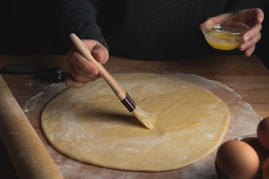 A man brushing dough with egg wash on a wooden table, showing the process of preparing pastry.