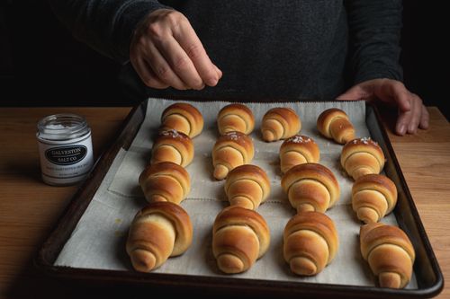 Free A person sprinkles sea salt on golden croissants, freshly baked on a tray. Stock Photo