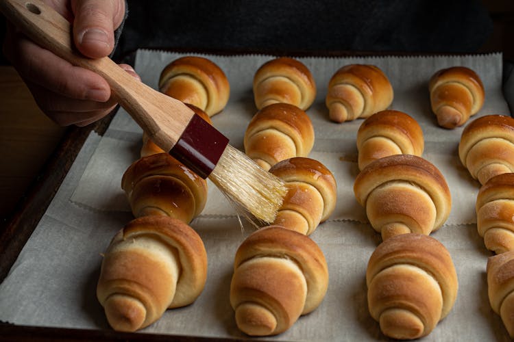 A Person Applying Oil On Croissants With A Wooden Brush