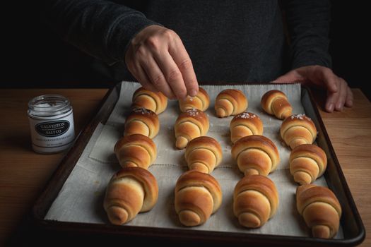 Artisan baker seasoning freshly baked croissants with sea salt on a tray.