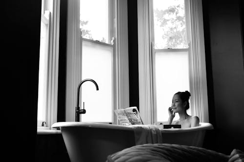Grayscale Photo of a Woman Holding a Book in a Bathtub