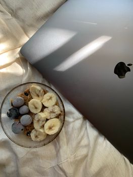 Overhead shot of a breakfast bowl with bananas and blueberries next to a laptop on a bed.