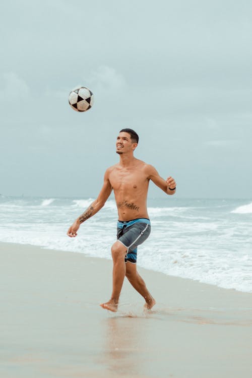 A Man in Blue Shorts Playing Soccer on the Beach