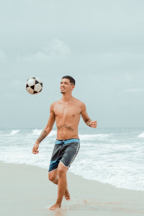 Photo of a Shirtless Man Playing with a Soccer Ball at the Beach