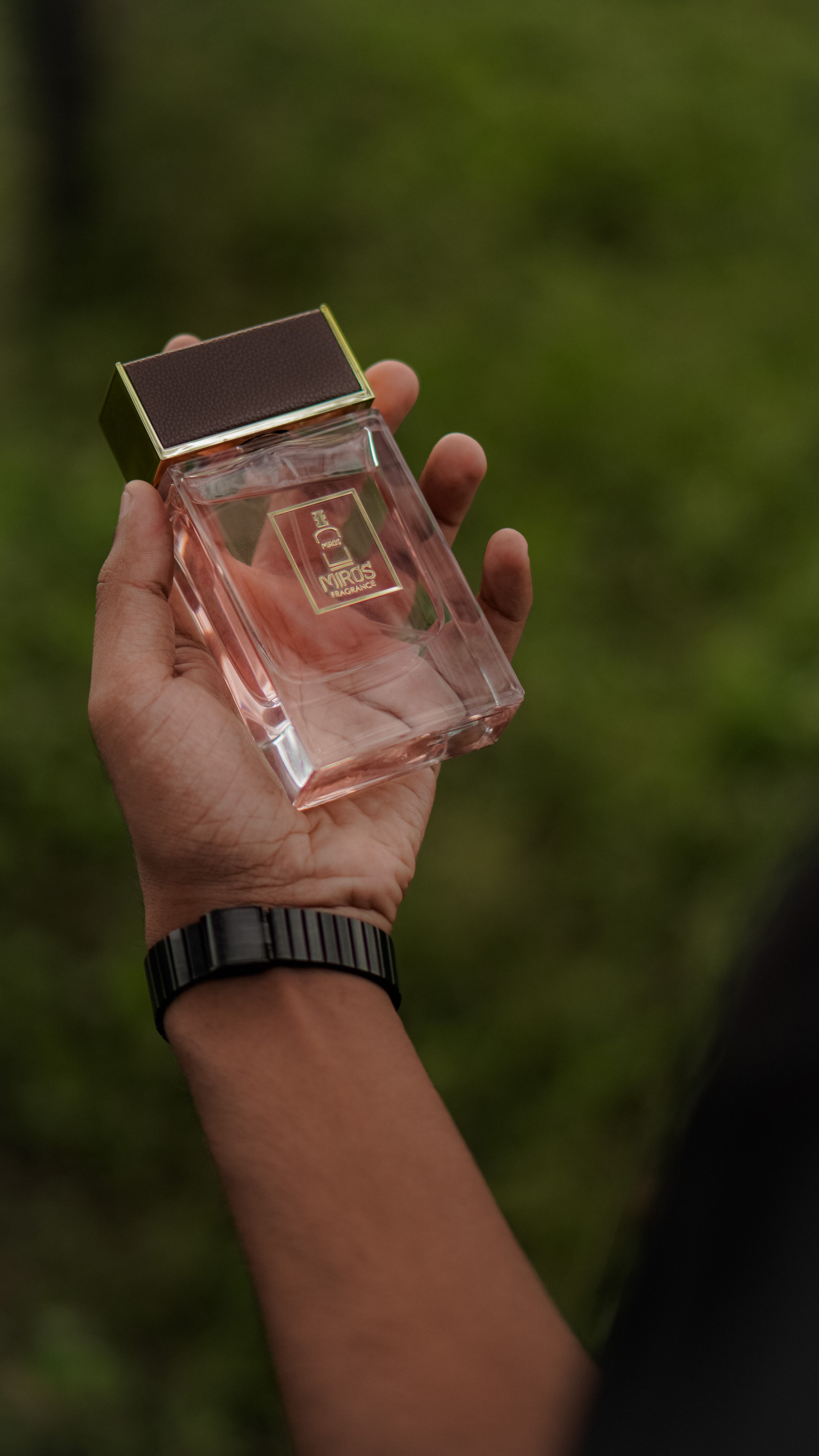 Close-up de uma mão segurando um frasco de perfume de luxo contra um fundo verde desfocado, mostrando elegância.