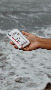 A smartphone held by a hand with a Paris luggage tag, against a beach backdrop.