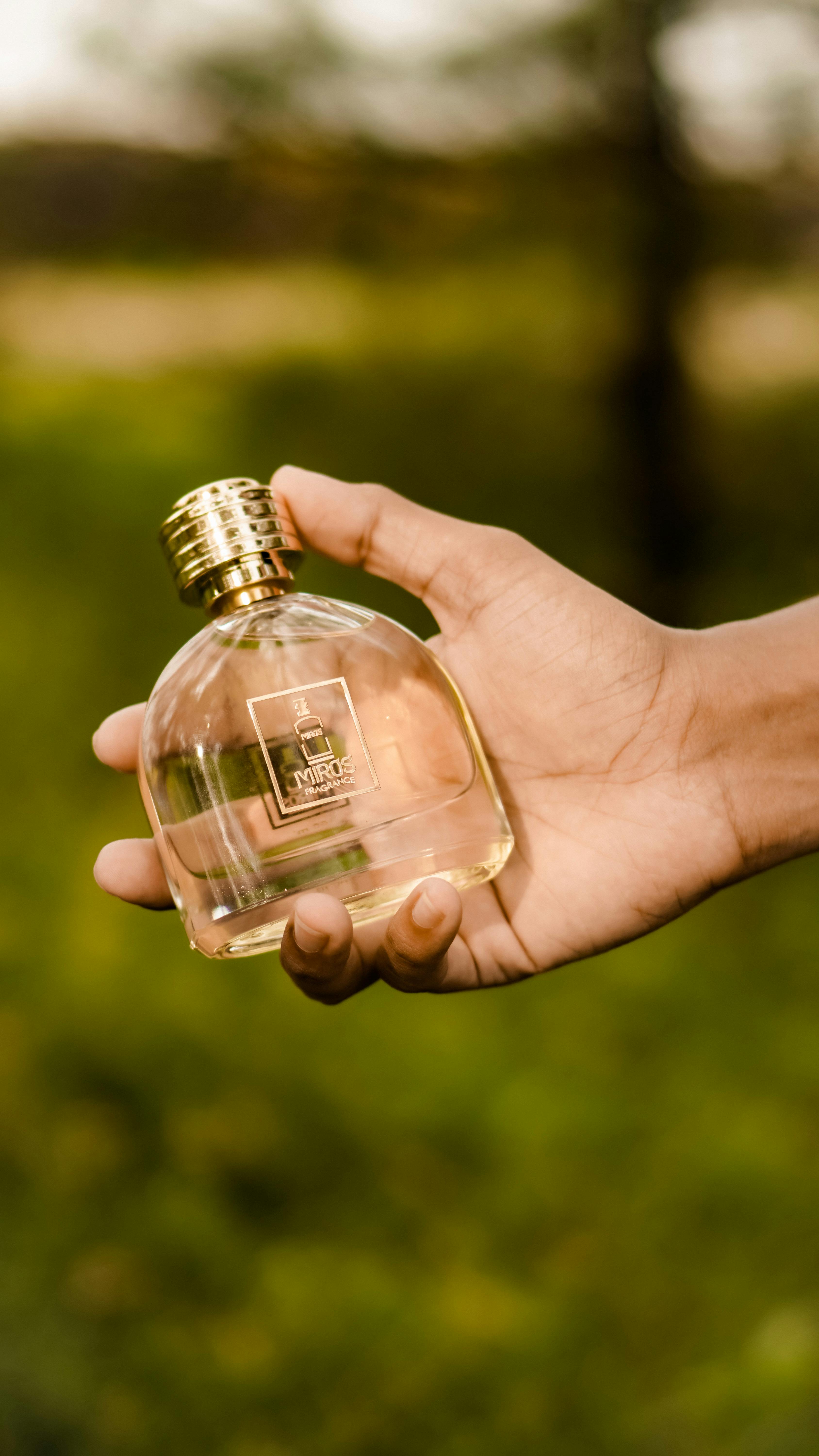 A clear glass perfume bottle with a gold cap held by a hand in an outdoor setting.