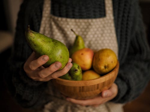A Person Holding Green Pear and a Wooden Bowl Filed with Fruits