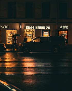 Night street view with illuminated cafes and cars reflecting on wet pavement.