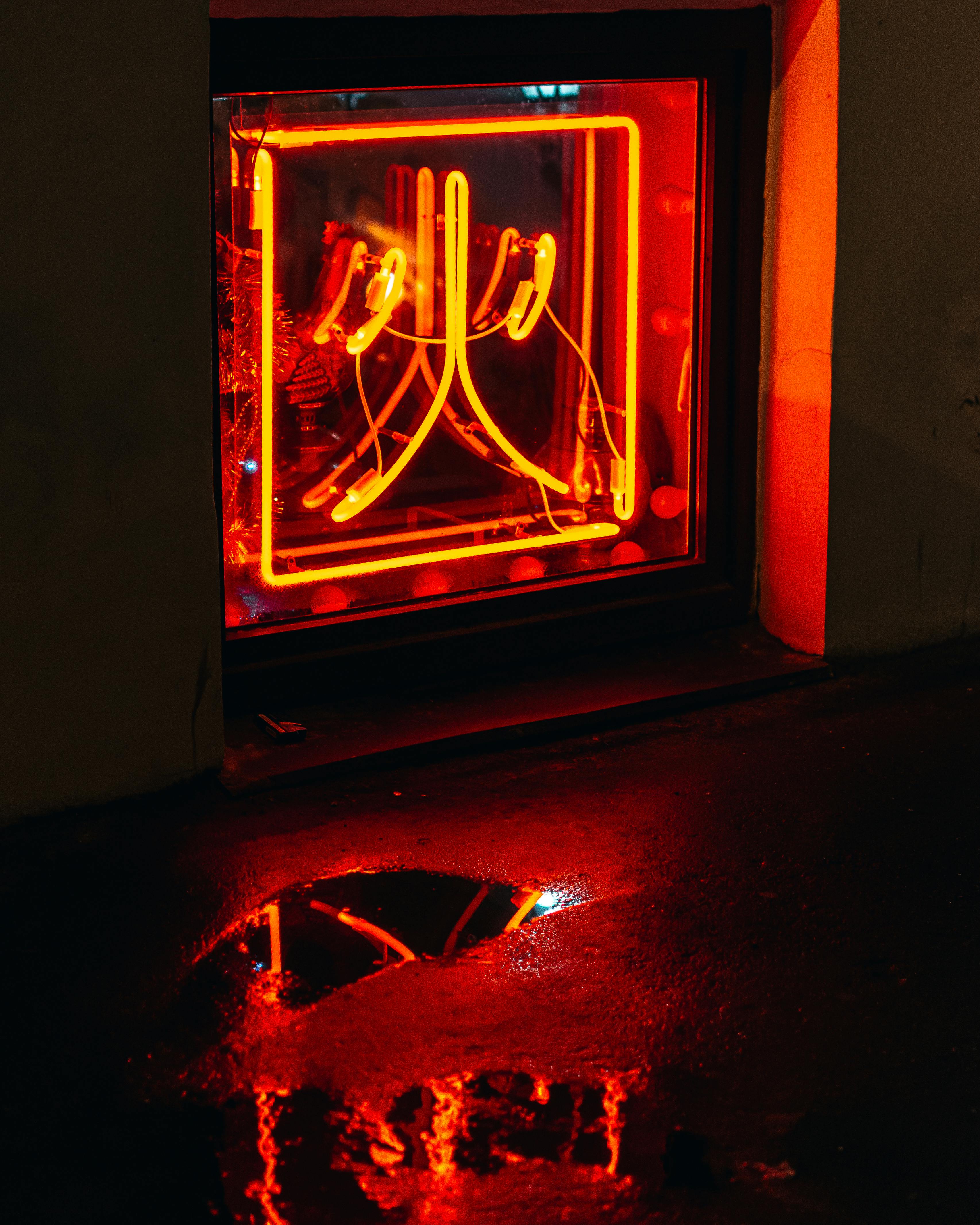 A close-up of a bright neon sign with its reflection on a wet street at night, creating a moody urban scene.