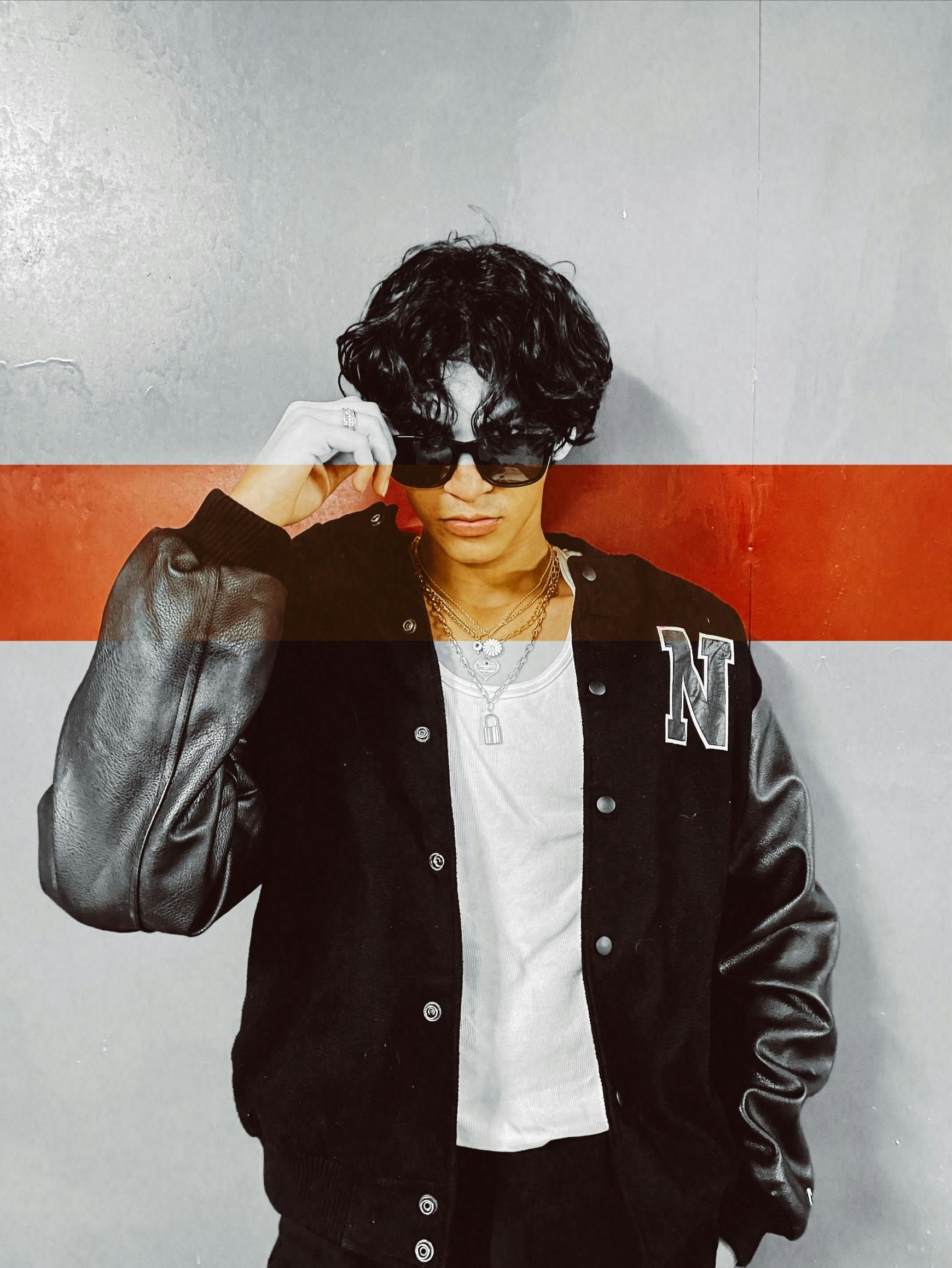 Fashionable young man poses in black varsity jacket and sunglasses indoors with a confident stance.