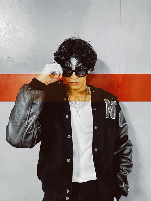 Fashionable young man poses in black varsity jacket and sunglasses indoors with a confident stance.