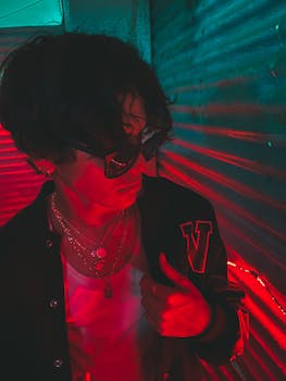 Man in black jacket with sunglasses and necklaces in vibrant neon red and green light.
