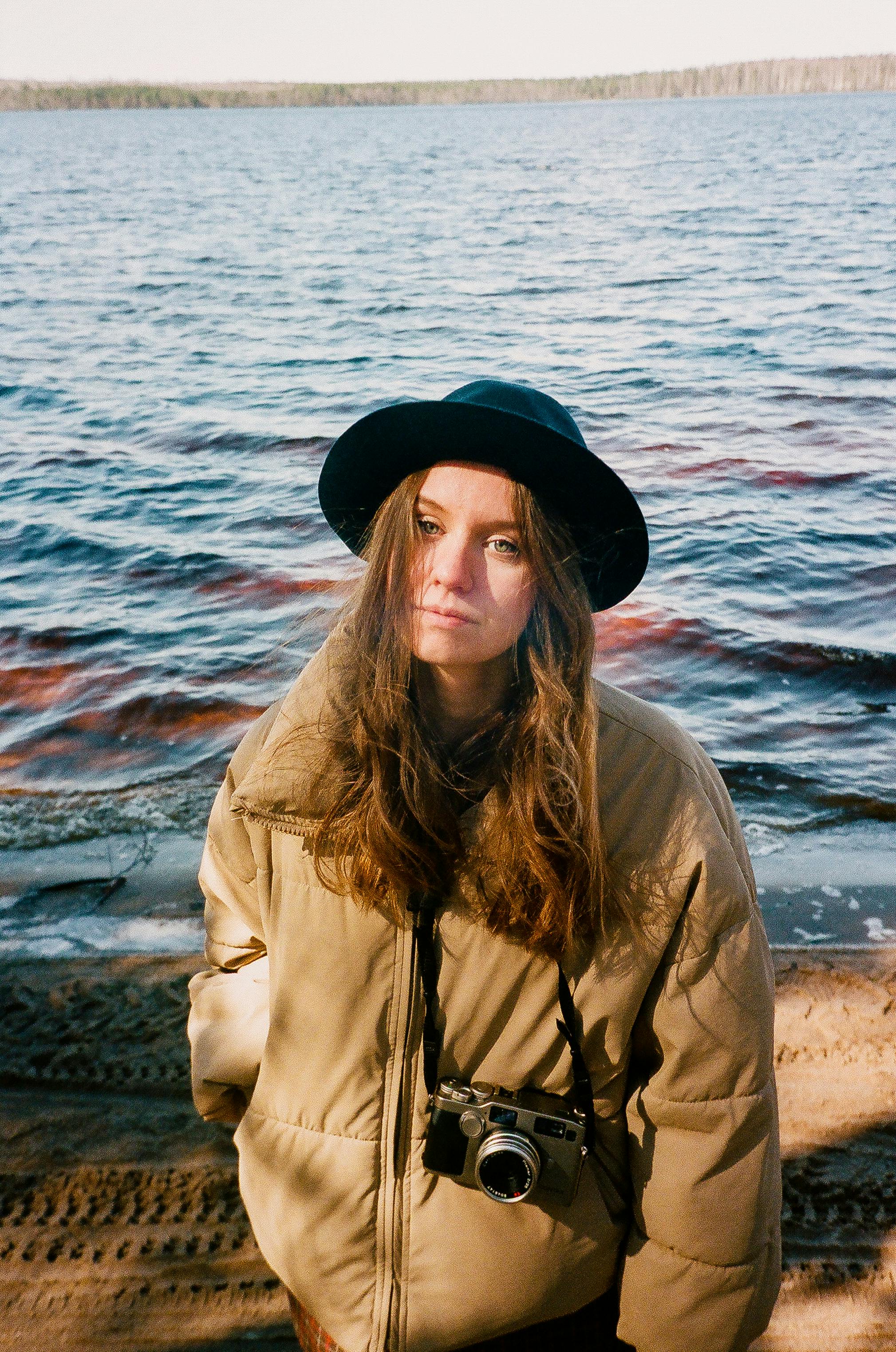 Young woman with a camera and hat poses by a serene lake on a sunny day.