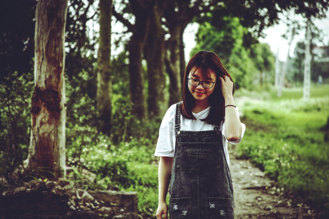 Free Woman Wearing White V-neck T-shirt and Black Denim Dungaree Standing Beside Trees Stock Photo