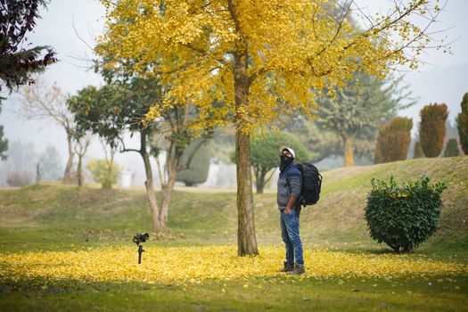 A man with a backpack stands under a tree with yellow leaves in a serene autumn park.