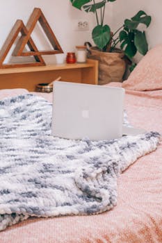 A cozy bedroom setup with a laptop on a bed covered by a plush blanket.