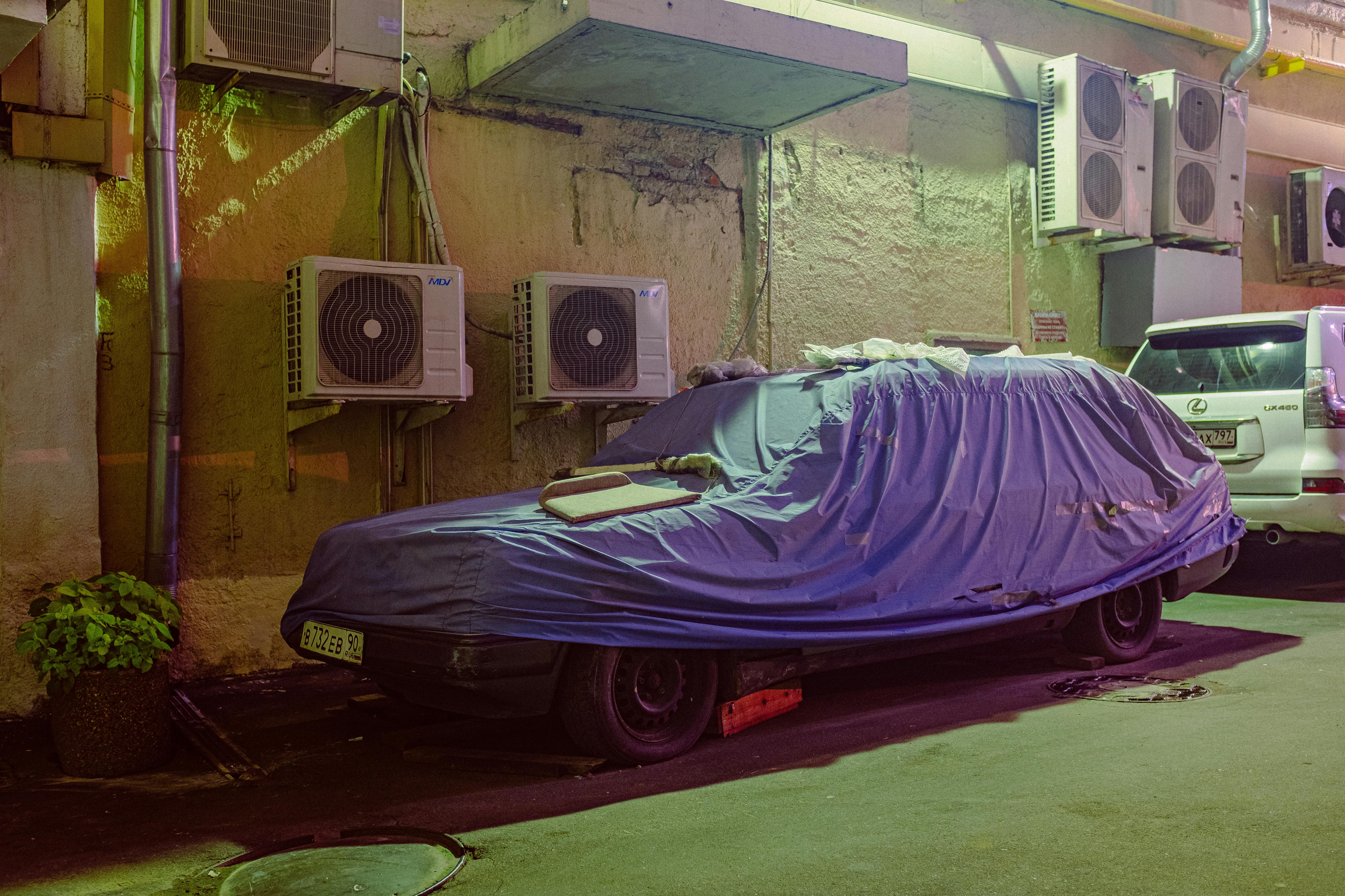 A car covered with a protective sheet parked in an urban alleyway surrounded by air conditioning units.