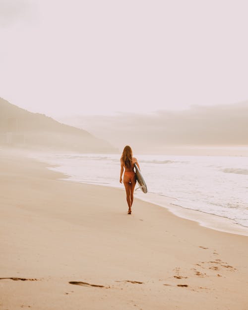 A Shot From Behind of Female Walking a Coastline With Surfboard