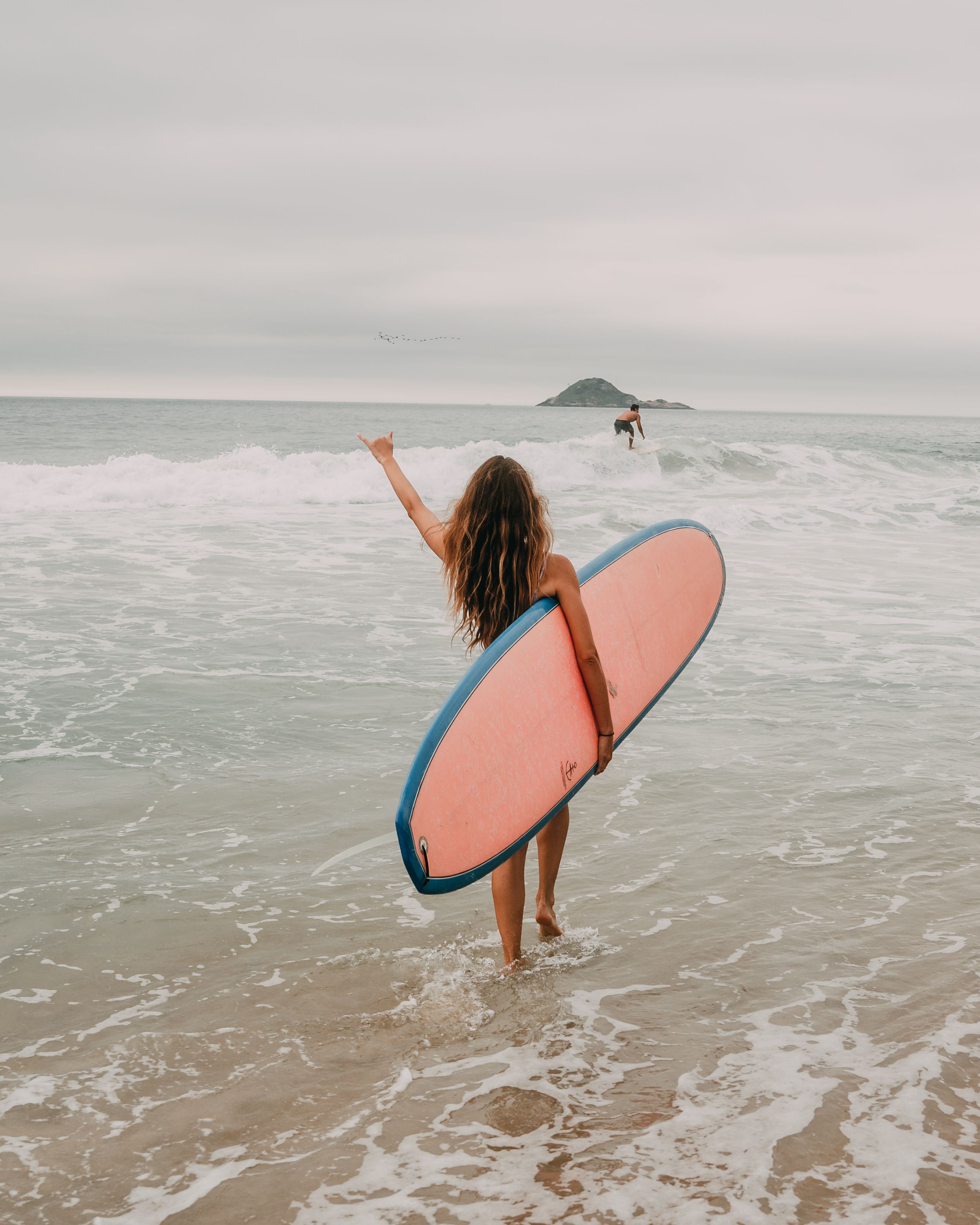 A woman with a surfboard walks towards the ocean waves at a beach in Rio de Janeiro, Brazil.